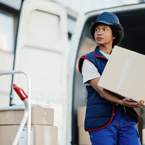 Black female courier unloading cardboard boxes from a van while making a delivery in the city.