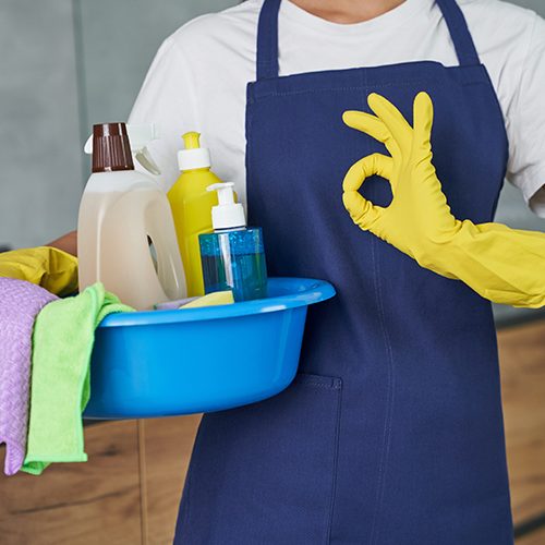 Cropped shot of cleaning lady showing ok sign while holding container full of cleaning products and equipment, standing in the modern kitchen. Housework and housekeeping, cleaning service concept